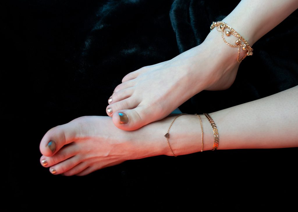Crop anonymous pedicured female legs in precious golden anklets lying on black background